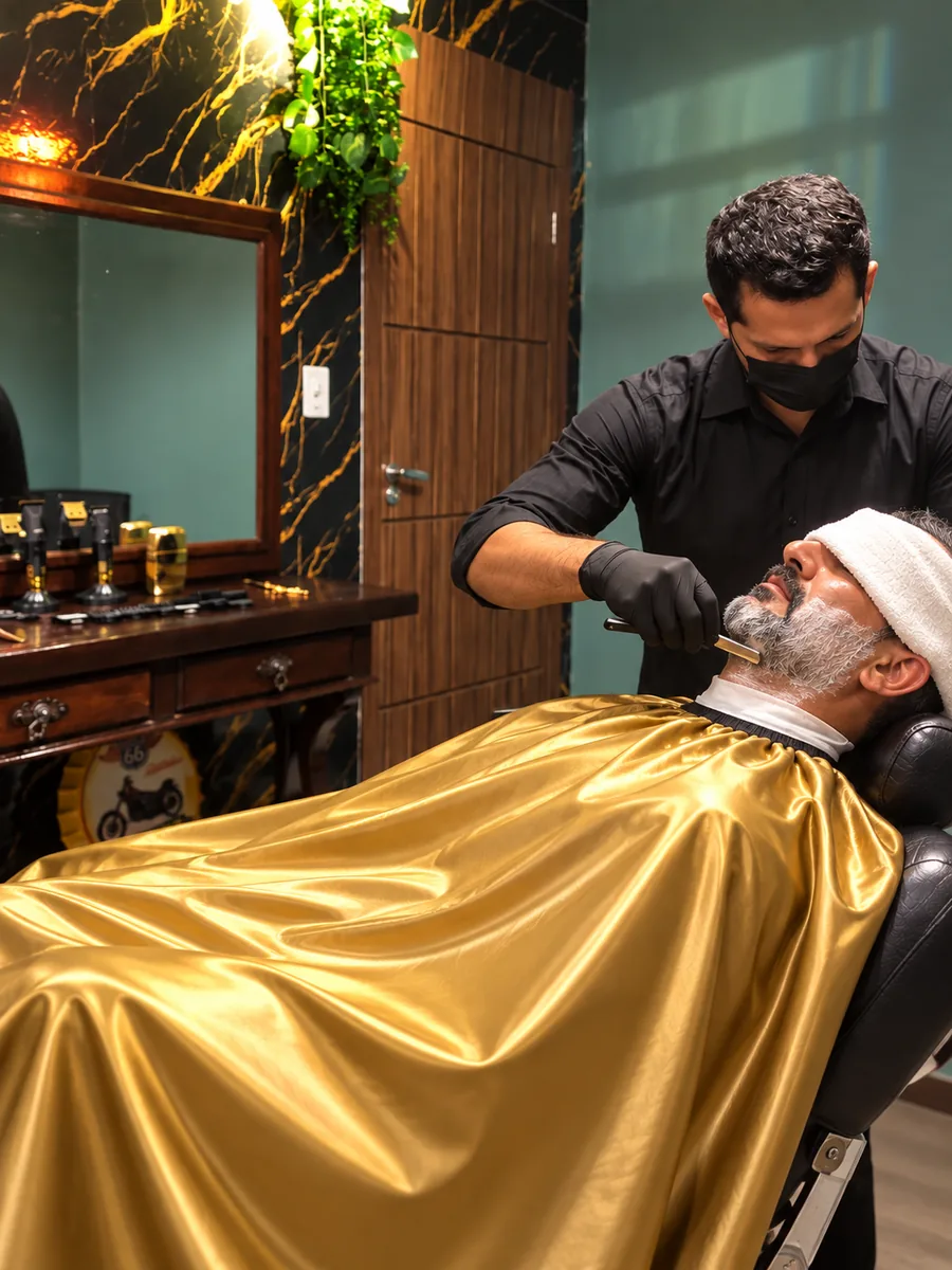 Barbeiro fazendo a barba com navalha em cliente reclinado na cadeira, coberto por capa dourada, com bancada de madeira e parede em mármore preto com veios dourados